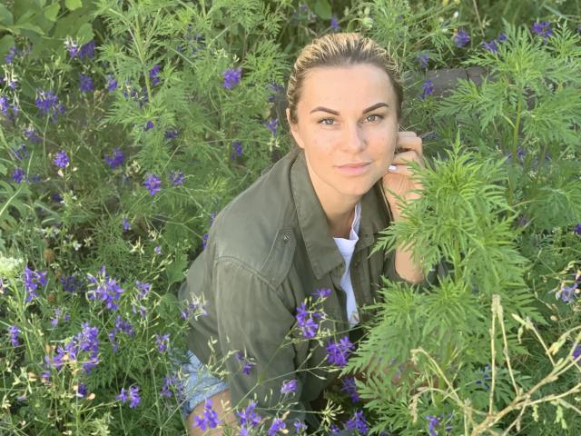 BelleLisaG with green eyes, medium tits, and light brown hair in a green military-style shirt, kneeling among purple flowers, greenery, and ferns, smiling softly at the camera.
