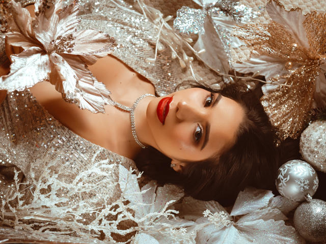 Il viso di Bellasweeth, con rossetto rosso, capelli scuri e una collana di perle, giace su pizzo argentato e dorato, circondata da ornamenti scintillanti, e guarda verso l'alto con un'espressione sexy e seducente..