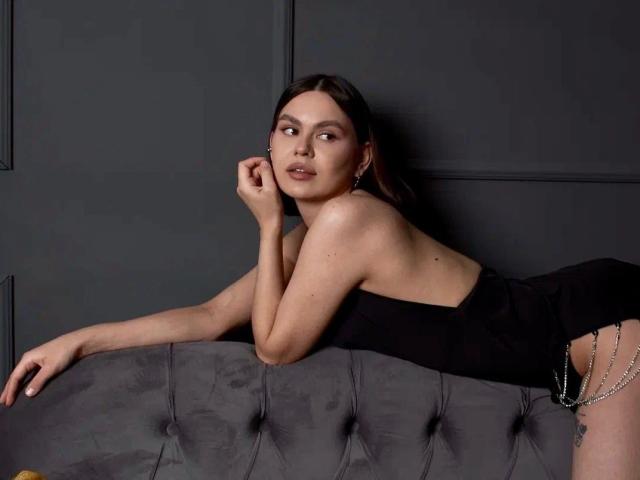 Sextenia-hot leans on a gray tufted sofa. She wears a black strapless dress with a chain accent on the right. Her right arm rests on the sofa, left hand near her chin. She has light skin, brown hair, and subtle makeup with pink eyeshadow. She gazes slightly to the right. The image uses soft lighting, highlighting her relaxed pose and the dress's texture.