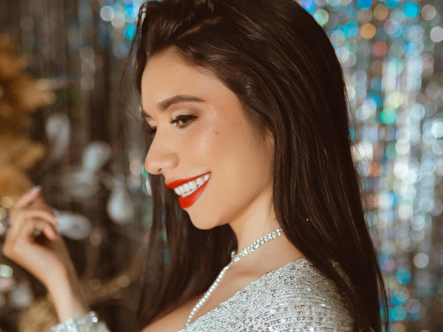 Bellasweeth heeft glanzend bruin haar, rode lippenstift en een parelketting, en ze glimlacht terwijl haar borsten licht zichtbaar zijn onder een zilveren top. Ze houdt haar vinger omhoog..