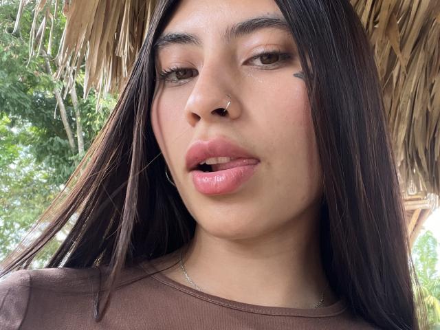 Macarenaa69 with long brown hair, nose ring, pink lips, slightly parted, brown eyes, subtle makeup, brown shirt, green tattoo on right cheek, under a straw roof.