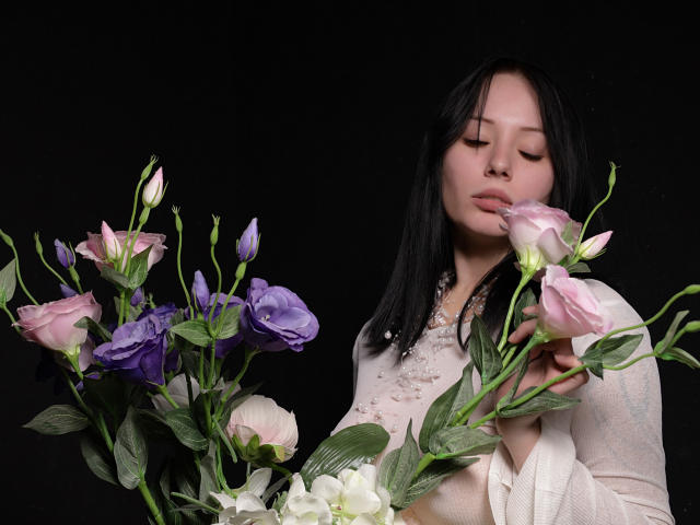 EliseVance with black hair, eyes closed, holding pink and purple roses, wearing a white sheer top, against a black background. Her tits are partially visible under the top.