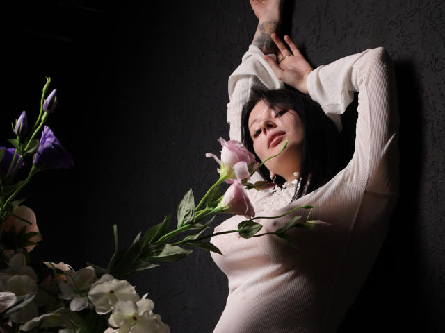 EliseVance with arms above head, wearing a white long-sleeve top, holding pink and purple flowers, tattoo on hand, dark short hair, soft light, black textured wall.