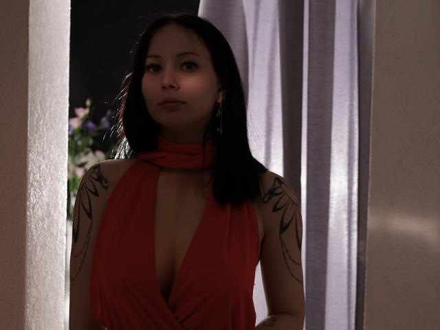 EliseVance with black hair, tattoos on her arms, wearing a red halter dress with deep cleavage, standing in a doorway, white curtain in background, looking serious.