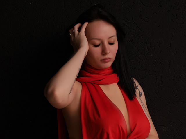 EliseVance with black hair, pale skin, and a red scarf, has her hand on her head, showing a tattoo on her arm, with her tits barely covered.