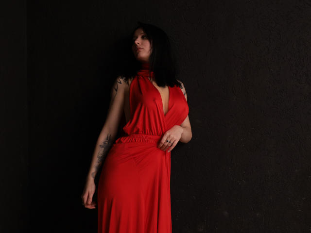 EliseVance with black hair, red dress, visible tattoos, standing against dark wall, right hand on waist, left arm relaxed, looking up, dark shadows, red dress highlights her tits.