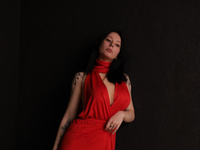 EliseVance with black hair, red dress, deep cleavage, tattoos on arms, leaning against dark wall, looking seductively at camera, hand on hip, dark lighting, soft shadows.