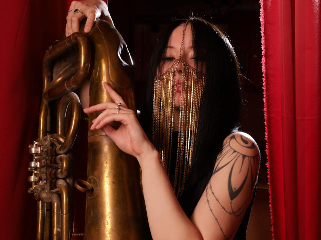 EliseVance with long black hair, wearing a gold chain mask, holds a brass pipe. She has a tattoo on her right arm, and red curtains are in the background.