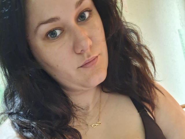 LoreneKisa has wavy dark brown hair, blue eyes, a nose ring, and a gold infinity necklace. She's wearing a black strap top, showing her tits.