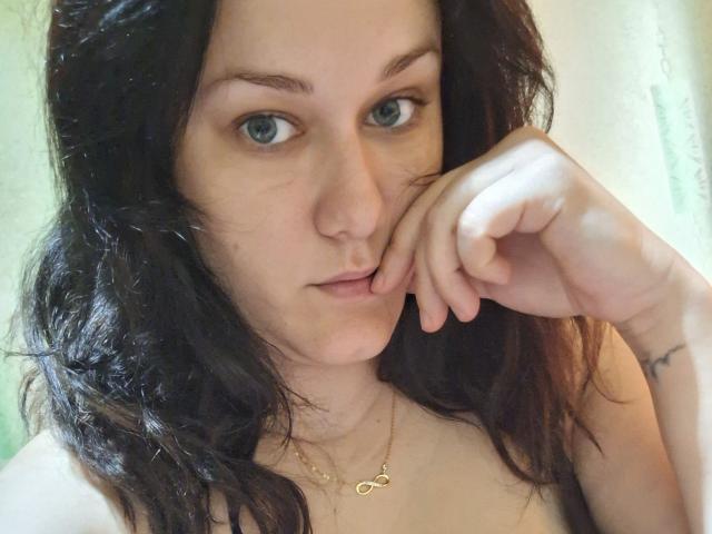 LoreneKisa has wavy dark hair, blue eyes, and a slight pout with her finger touching her lower lip, wearing a gold necklace with a small heart.