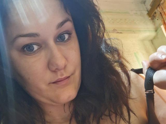 LoreneKisa with wavy brown hair, blue eyes, and a slight smile, holding a black strap, looking at the camera with a relaxed, confident expression.