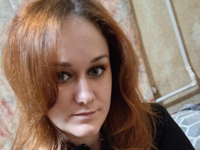 LoreneKisa has red hair, light skin, and blue eyes. She's wearing a black top, with a subtle smile and looking directly at the camera.