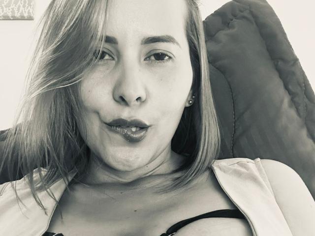 Sarita Velez, com seios grandes e cobertos por rendas, usando um sutiã preto e rosa, uma camisa bege puxada para baixo, cabelo loiro e um sorriso discreto, sentada em um quarto claro..