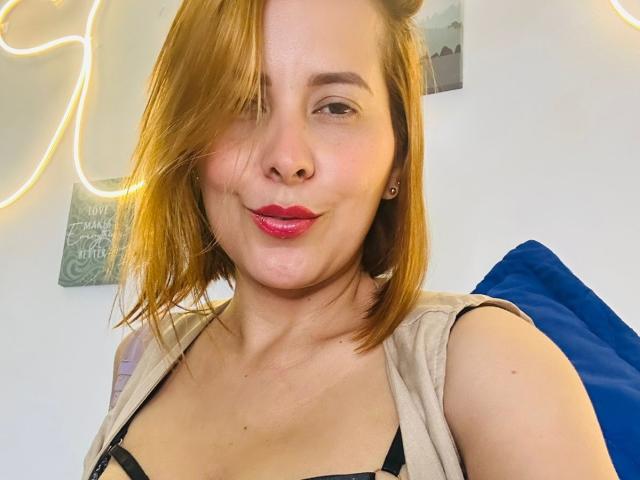 SaritaVelez, a young woman with fair skin and shoulder-length red-brown hair, takes a selfie. She has a slight smile with pink lipstick and a small nose piercing. She wears a beige sleeveless top over a black strap, exposing her cleavage. The background includes a blue pillow and a white wall with a neon light outline and a green wall art piece. Her hair is partially covering her right eye. The image is clear and focused on her upper body.