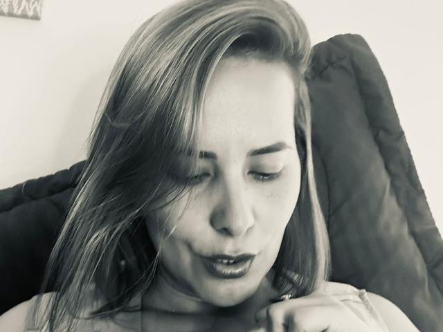 SaritaVelez with puffy lips, blonde hair, and a light tan shirt open to show black and lace-patterned bra covering her big tits. Neon light in the background.
