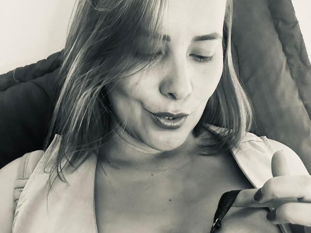 SaritaVelez hat langes, leicht zerzaustes Haar, halb geschlossene Augen und dunkelroten Lippenstift, mit einem entspannten, leicht verschmitzten Ausdruck, und sitzt vor einem dunklen, strukturierten Hintergrund..