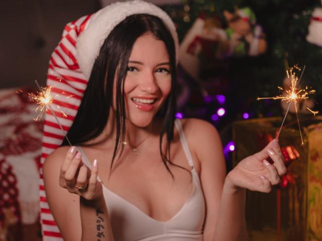 LizzeJoy with long black hair, wearing a Santa hat, white tank top, smiling, holding sparklers, showing her small tits, tattoo on right arm, in a Christmas-themed setting.
