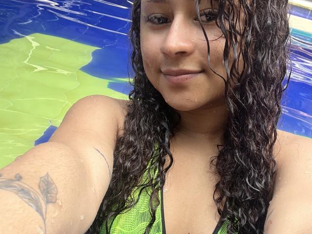 SofiaPassionne with wet, curly dark hair, tan skin, green swimsuit, small tattoo on right arm, smiling at camera, water droplets on face and chest. Boobs visible through swimsuit.