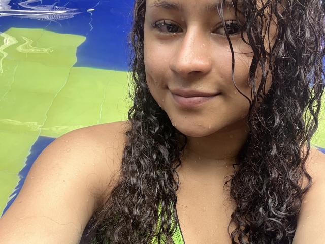 SofiaPassionne has curly wet brown hair, tan skin, and small nose, wearing a green bikini top, smiling slightly, with water droplets on her face and shoulders.