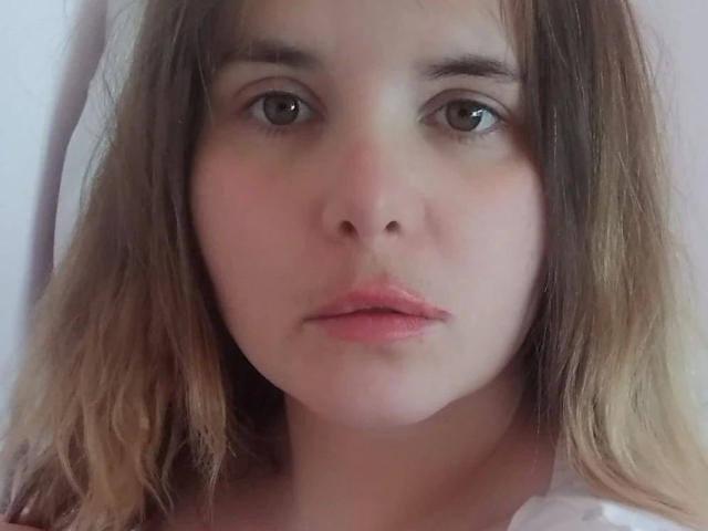 Close-up of ElenaSchatz-hot with a neutral expression. She has light skin, brown eyes, and slightly parted pink lips. Her brown hair is wavy and falls to the sides, with lighter ends. The image is centered on her face, showing soft, natural light. The background is blurred, emphasizing her facial features. The texture of her hair is slightly messy, and she wears a white garment. The image has a candid, intimate feel.