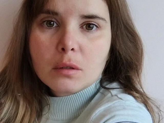 ElenaSchatz-hot has pale skin, brown eyes, and wavy brown hair, wearing a light blue turtleneck. Her expression is neutral, with slightly parted pink lips.