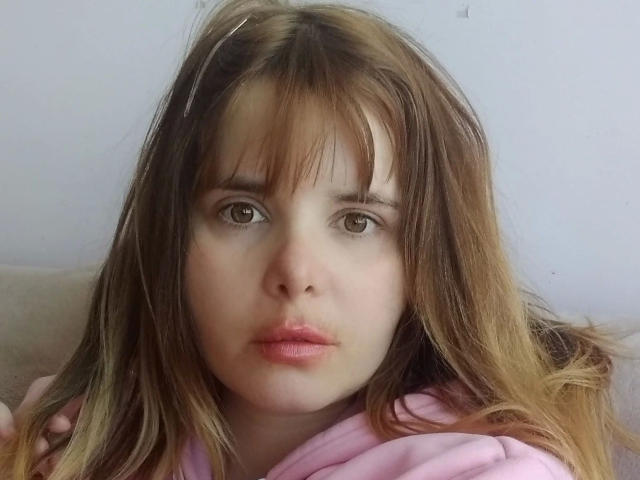 ElenaSchatz-hot has brown eyes, pink lips, and messy brown hair with bangs, wearing a pink hoodie, looking serious. Her face is centered, with a neutral background.