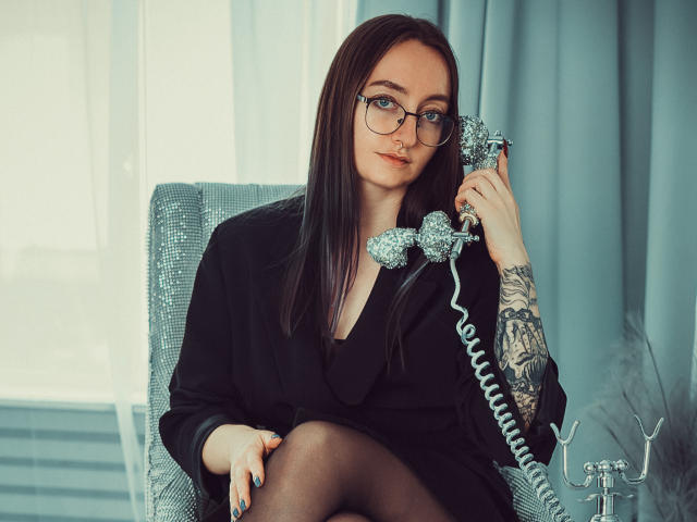 Shiroheby con gafas, vestido negro y medias negras, sosteniendo un teléfono de plata antiguo con un cable enrollado, sentada en una silla gris, mostrando un tatuaje en su mano derecha..