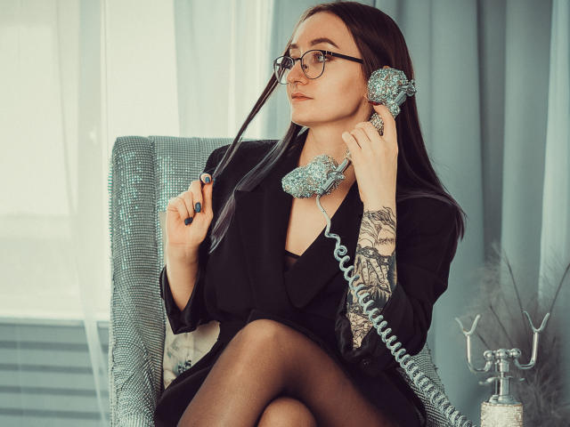 Shiroheby con gafas, sosteniendo un teléfono azul con flores pegadas cerca de su oreja, sentada en una silla azul, vistiendo un blazer negro, un vestido negro y medias de nylon transparentes, con tatuajes en su brazo..