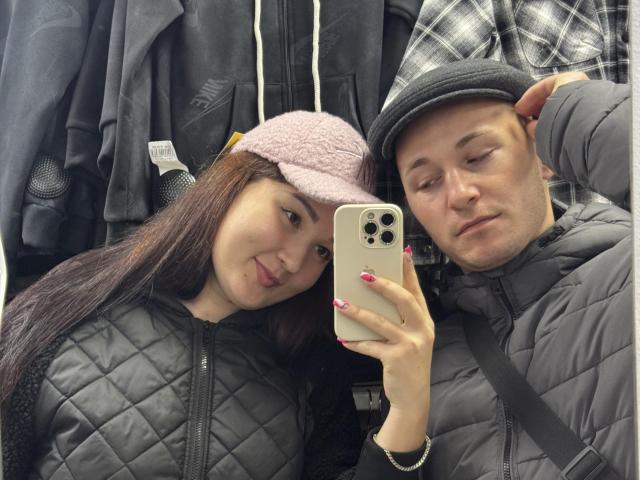 DianaAndErnest have a selfie, she's wearing a pink fuzzy hat and a black quilted jacket, he's in a gray flat cap and gray jacket, holding a white iPhone.