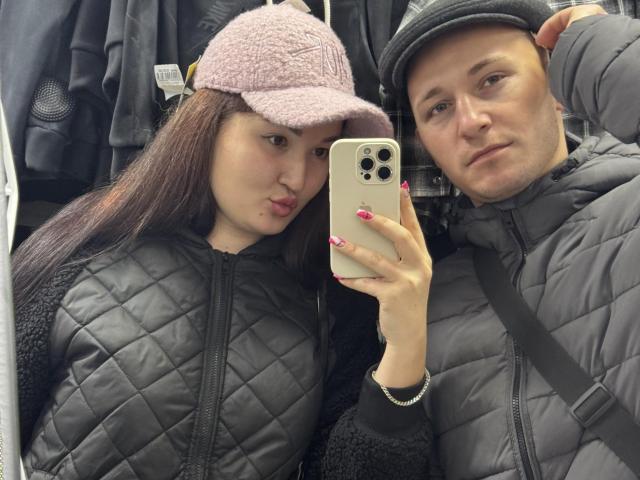 DianaAndErnest with pink fuzzy cap, black quilted jacket, pink nails, holding phone, standing next to Ernest in gray quilted jacket, black cap, mirror selfie, store background.