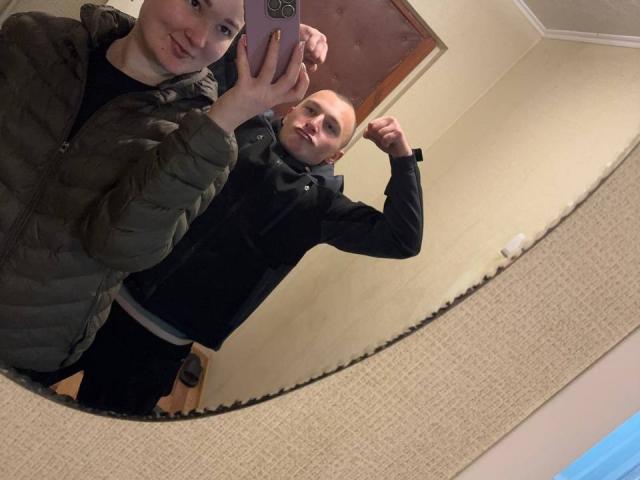 DianaAndErnest with pale skin, green jacket, and black shirt, taking a mirror selfie. Ernest, bald, black jacket, flexing bicep, stands behind. Both smiling. Bedroom setting.