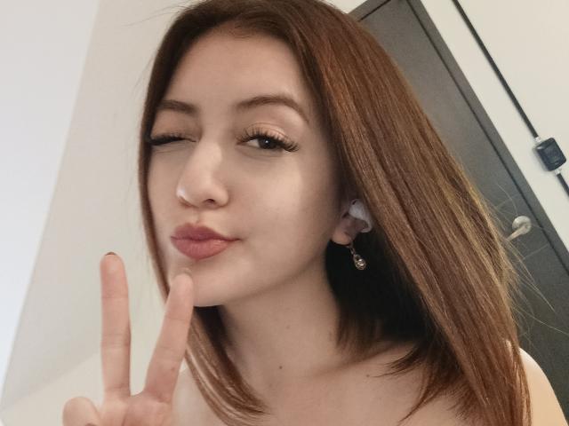 AngelaTorres with brown hair, winking, making a peace sign, wearing a nose ring and ear cuff, pink lips, white skin, subtle makeup, looking cute, showing off her tits.