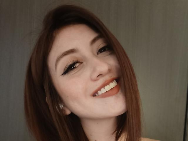 AngelaTorres has light skin, brown hair, black eyeliner, and a smile showing white teeth. She has a small mole under her right eye and is looking up.