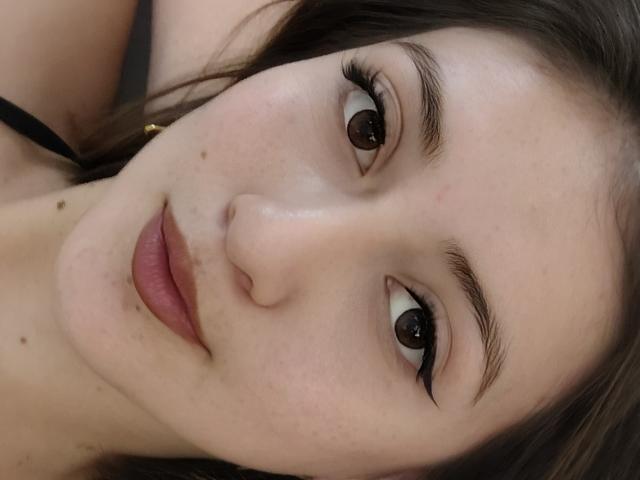 AngelaTorres has light skin, brown eyes, and dark brown hair. She's looking at the camera with a slight smile, showing small moles on her cheek and nose.