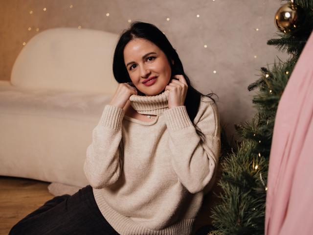 TayaBoret, con lunghi capelli neri, seduta a terra, indossa un maglione beige a collo alto, le mani vicino al collo, sorride, un albero di Natale a destra, un tessuto rosa a sinistra, pavimento in legno..
