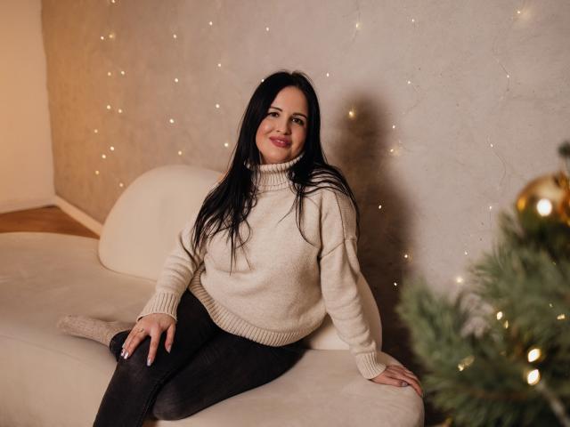 TayaBoret with long black hair, smiling, wearing a beige turtleneck sweater, black pants, sitting on a white couch, Christmas tree with ornaments in the blurred foreground.