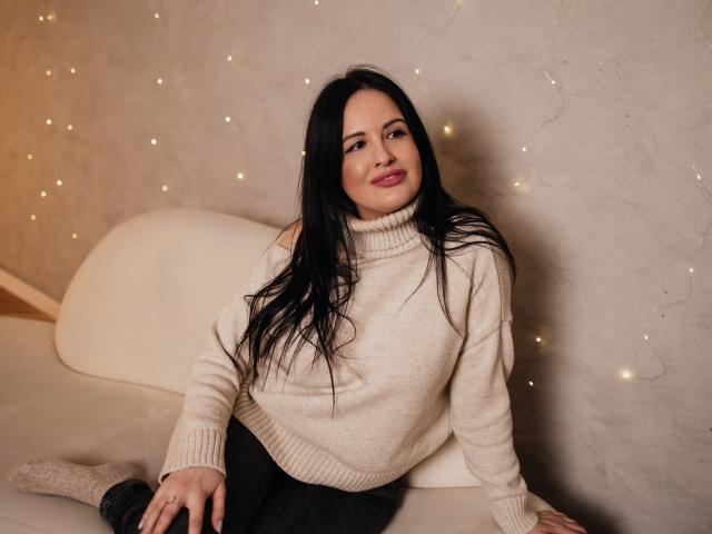 TayaBoret with long black hair, red lipstick, and a white turtleneck sweater, sitting on a beige couch, looking at the camera with a slight smile, wearing black pants.
