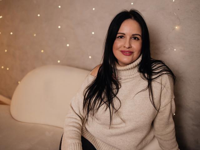 TayaBoret with long black hair, smiling, wearing a beige turtleneck sweater, sitting on a white couch, her small tits slightly visible under the sweater.
