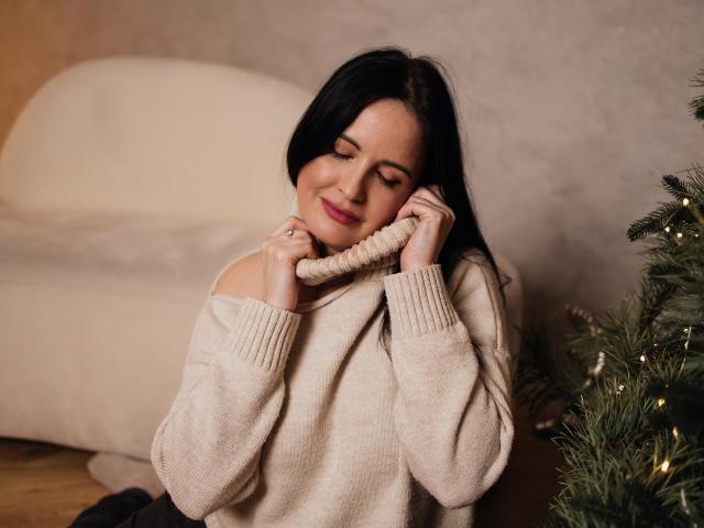 TayaBoret, com cabelo preto, olhos fechados e batom rosa, vestindo um suéter de malha bege com um ombro descoberto, segurando um cachecol de malha no rosto, sorrindo. Árvore de Natal à direita..