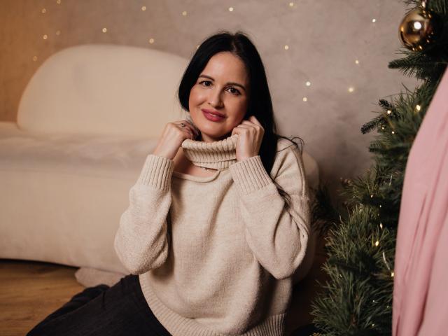 TayaBoret with long black hair, smiling, holding up a beige sweater, wearing a black skirt, sitting by a Christmas tree, wood floor, soft pink couch, warm lighting.