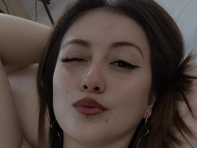 AngelaTorres with one eye closed, brown eyeliner, red lipstick, freckles, and small moles, winking, wearing hoop earrings, lying down, and looking at the camera.