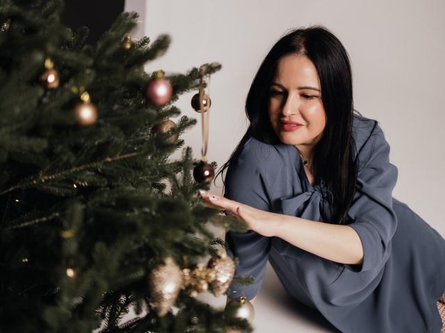 TayaBoret with black hair, wearing a blue shirt, kneeling by a Christmas tree, gently touching a golden ornament, smiling, looking at it with a calm, satisfied expression.