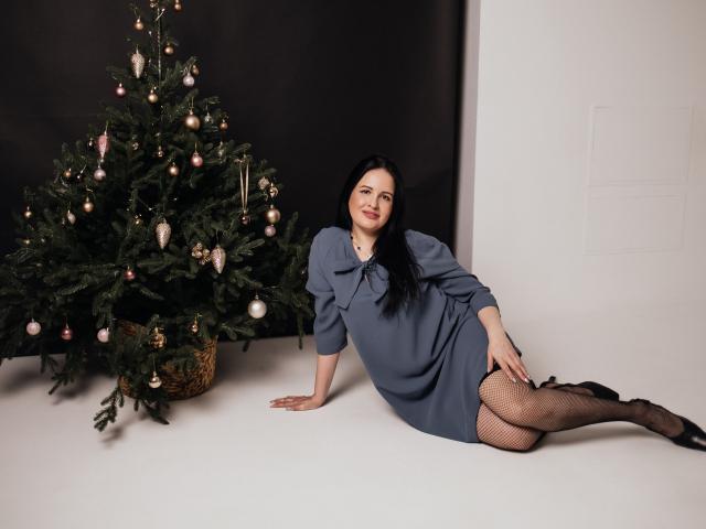TayaBoret, com cabelo preto, vestido azul e meias de rede, deitada no chão branco, com os braços atrás, ao lado de uma árvore de Natal decorada. Seios visíveis sob o vestido..