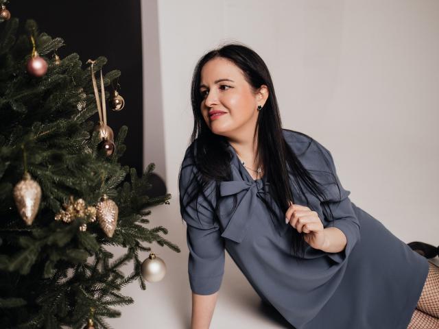 TayaBoret, com longos cabelos pretos, vestindo um vestido azul e brincos, apoiada em uma árvore de Natal decorada, sorrindo e segurando o laço do vestido. Seios não visíveis..
