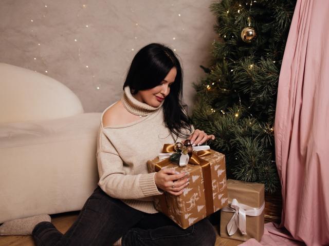TayaBoret, com cabelo preto, vestindo um suéter bege com um ombro descoberto e calças escuras, sentada perto de uma árvore de Natal, sorrindo para um presente embrulhado em dourado e marrom, com um laço branco..