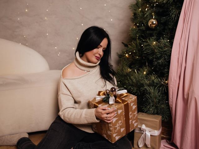 TayaBoret with black hair, off-shoulder beige sweater, and dark pants, sitting by a Christmas tree, holding a gold-wrapped gift with a brown ribbon.