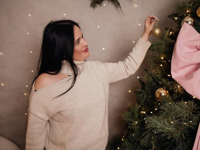 TayaBoret with long black hair, off-shoulder beige sweater, hanging gold ornament from Christmas tree, pink ribbon in background, warm fairy lights, soft smile, elegant and cozy.