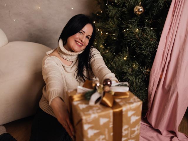 TayaBoret, com cabelo preto, sorrindo, vestindo um suéter de gola alta bege, segurando um presente embrulhado em papel dourado com um laço, ao lado de uma árvore de Natal com luzes..