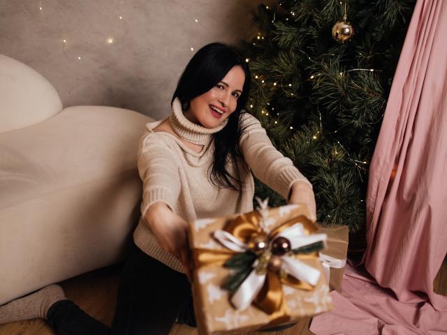 TayaBoret with black hair, smiling, wearing a beige sweater, kneeling, holding a gold-wrapped gift with a ribbon, in front of a Christmas tree. Boobs not visible.