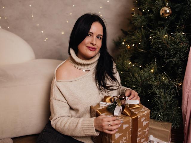 TayaBoret, com cabelo preto, vestindo um suéter de gola alta bege, segurando um presente embrulhado em papel dourado com uma fita dourada, sentada ao lado de uma árvore de Natal com pequenas luzes..